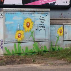 Stromkasten an einer Hauswand, besprüht mit gelb blühenden Blumen und dem Schriftzug "Support your local Antifa".