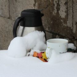Kaffeekanne und zwei Tassen im Schnee auf einem kleinen Tisch.